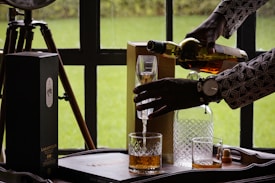 A person wearing a patterned shirt is pouring a glass of whiskey or a similar beverage from a bottle. The scene includes a wooden table or tray with a box labeled 'Lagavulin 16', a couple of glasses, one with liquid and the other empty, a glass decanter, and a wristwatch visible on the person's wrist. The background shows a green outdoor area through a window.
