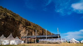 A large outdoor venue set against a rocky cliff with several white tents and a stage equipped with lighting and colorful decorations. The sky is clear and vibrant blue with some clouds.