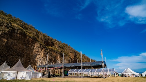 A large outdoor venue set against a rocky cliff with several white tents and a stage equipped with lighting and colorful decorations. The sky is clear and vibrant blue with some clouds.