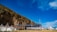 A large outdoor venue set against a rocky cliff with several white tents and a stage equipped with lighting and colorful decorations. The sky is clear and vibrant blue with some clouds.