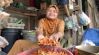 A woman in a brown hijab and embroidered clothing is sitting in a rustic marketplace. She is smiling and holding out a handful of vibrant red and green chili peppers. Around her, there are various items like plastic bags containing produce, baskets, and packaged goods in a wooden stall.