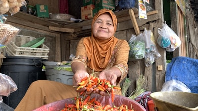 A woman in a brown hijab and embroidered clothing is sitting in a rustic marketplace. She is smiling and holding out a handful of vibrant red and green chili peppers. Around her, there are various items like plastic bags containing produce, baskets, and packaged goods in a wooden stall.