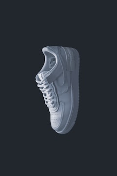 A single white sneaker is prominently displayed against a dark background. The shoe has a clean and minimal design with visible stitching, a perforated toe box, a lace-up closure, and a distinct sole. The lighting casts subtle shadows, emphasizing the contours and texture of the sneaker.