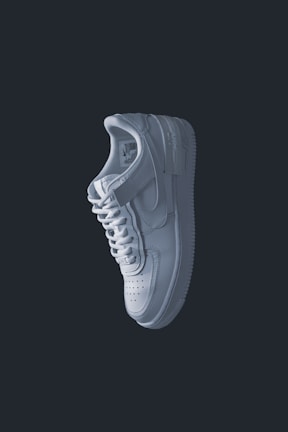 A single white sneaker is prominently displayed against a dark background. The shoe has a clean and minimal design with visible stitching, a perforated toe box, a lace-up closure, and a distinct sole. The lighting casts subtle shadows, emphasizing the contours and texture of the sneaker.
