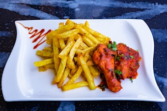 A white rectangular plate holds a serving of golden French fries arranged next to two pieces of red, spicy chicken garnished with herbs. A zigzag of sauce is artistically drizzled on the side of the plate, set against a dark, speckled background.