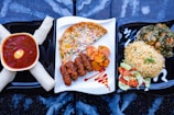 A visually appealing arrangement of African cuisine is presented on three plates. The left plate features a red soup with a boiled egg, surrounded by rolled white dough. The middle plate shows an omelette with colorful vegetables, grilled meat, and fried plantains with a decorative sauce drizzle. The right plate contains jollof rice with greens and chicken, accompanied by a salad of cucumbers and tomatoes.