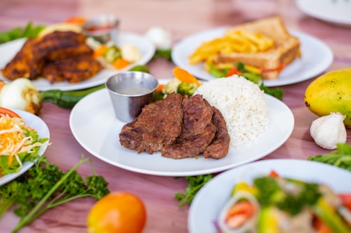 A white plate containing grilled meat slices, a serving of white rice, and a small metal cup of sauce is at the center. Surrounding the central plate are other dishes, including a plate with a sandwich and fries, a plate with fried plantains, a plate with mixed salad, and various fresh vegetables such as tomatoes, onion, and garlic placed on the table.