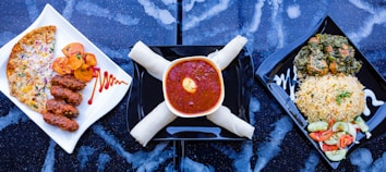 A trio of meals is arranged on a dark, speckled surface. On the left, a white square plate holds three pieces of grilled meat alongside slices of roasted potatoes and a cheese and vegetable-covered flatbread. The center shows a bowl filled with rich red stew topped with an egg yolk, accompanied by what appears to be rolled breadsticks. On the right, a square black plate contains a serving of rice garnished with herbs next to a mixed vegetable salad and a portion of leafy greens with meat.