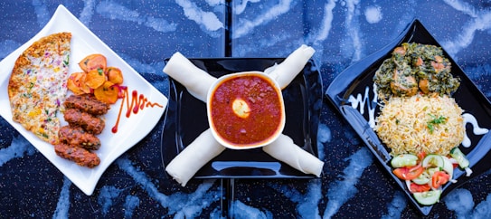 A trio of meals is arranged on a dark, speckled surface. On the left, a white square plate holds three pieces of grilled meat alongside slices of roasted potatoes and a cheese and vegetable-covered flatbread. The center shows a bowl filled with rich red stew topped with an egg yolk, accompanied by what appears to be rolled breadsticks. On the right, a square black plate contains a serving of rice garnished with herbs next to a mixed vegetable salad and a portion of leafy greens with meat.