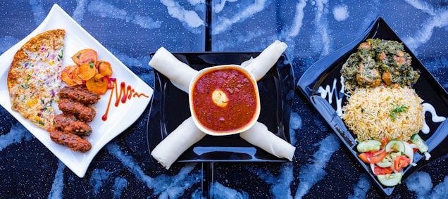 A trio of meals is arranged on a dark, speckled surface. On the left, a white square plate holds three pieces of grilled meat alongside slices of roasted potatoes and a cheese and vegetable-covered flatbread. The center shows a bowl filled with rich red stew topped with an egg yolk, accompanied by what appears to be rolled breadsticks. On the right, a square black plate contains a serving of rice garnished with herbs next to a mixed vegetable salad and a portion of leafy greens with meat.