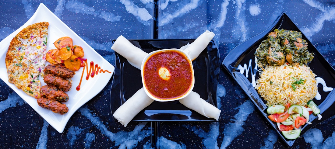 A trio of meals is arranged on a dark, speckled surface. On the left, a white square plate holds three pieces of grilled meat alongside slices of roasted potatoes and a cheese and vegetable-covered flatbread. The center shows a bowl filled with rich red stew topped with an egg yolk, accompanied by what appears to be rolled breadsticks. On the right, a square black plate contains a serving of rice garnished with herbs next to a mixed vegetable salad and a portion of leafy greens with meat.