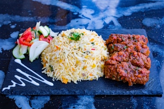 A serving of aromatic yellow rice garnished with herbs is paired with four pieces of spiced meat on a black rectangular slate. To the left, there is a side of fresh salad containing sliced cucumbers, tomatoes, and onions. A decorative swirl of white sauce is artistically placed on the slate.