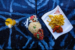 A dark marbled table surface holds a vibrant arrangement of food. On the left, there is a tall glass filled with a bright yellow smoothie, garnished with a slice of pineapple and a straw. Next to the drink, a black rectangular plate contains a portion of rice, garnished with herbs, alongside grilled meat in a red sauce, served with a creamy white sauce and salad made of cucumber and tomato slices. To the right, a white triangular plate holds golden french fries adorned with a zigzag of red sauce, accompanied by more pieces of grilled meat in a reddish marinade.