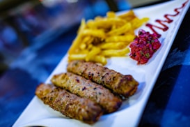 A white rectangular plate contains skewers of grilled meat, a serving of golden fries, a portion of chopped tomato salsa, and a drizzle of sauce.