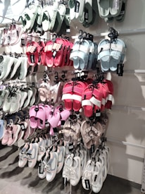 A variety of shoes are displayed on a wall-mounted rack, including sandals and sneakers in multiple colors, such as pastel green, pink, red, and white. Each pair is neatly hung on hooks, with visible brand tags.