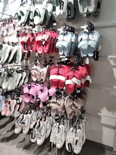 A variety of shoes are displayed on a wall-mounted rack, including sandals and sneakers in multiple colors, such as pastel green, pink, red, and white. Each pair is neatly hung on hooks, with visible brand tags.