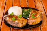 A plate of food featuring a serving of cooked ribs smothered in a rich sauce, accompanied by a portion of steamed greens and a mound of soft mashed potatoes or similar starchy dish, garnished with fresh parsley. The food is arranged neatly on a dark-colored plate placed on a wooden table that has a rustic look.
