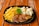 A dish consisting of a grilled steak covered in gravy, accompanied by seasoned French fries and a side of steamed vegetables including cauliflower, carrots, and corn. The meal is plated on a black plate and garnished with a sprig of parsley, placed on a wooden table.