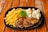 A dish consisting of a grilled steak covered in gravy, accompanied by seasoned French fries and a side of steamed vegetables including cauliflower, carrots, and corn. The meal is plated on a black plate and garnished with a sprig of parsley, placed on a wooden table.