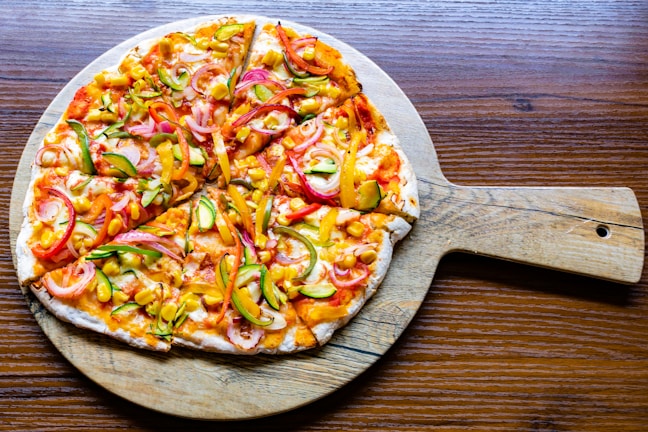 A colorful pizza topped with vibrant slices of red onion, yellow bell pepper, green zucchini, and corn pieces rests on a wooden pizza board. The crust appears crispy and lightly browned, while the toppings are arranged generously over a tomato-based sauce with melted cheese.