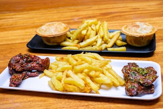 A wooden table features two rectangular platters filled with food. The foreground platter displays crispy golden fries and glazed chicken wings, garnished with herbs. The background platter contains two savory meat pies accompanied by another serving of fries.