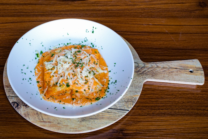 A round white plate on a wooden serving board holds a dish of ravioli covered in an orange cream sauce, garnished with grated cheese and finely chopped green herbs. The background is a wooden table surface.