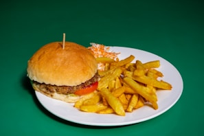 A large hamburger with a thick patty, lettuce, tomato, and sauce, placed on a white plate alongside golden fries and a small serving of coleslaw. The background is a solid green color.