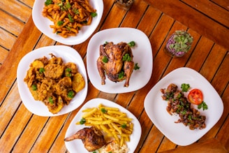 A rustic wooden table with a rotating spit of golden roasted chicken surrounded by smoked ribs and a bowl of saucy potatoes.