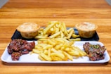 A wooden table holds a serving platter featuring crispy fries in the center, flanked by two small meat pies. Adjacent to the fries are portions of glazed chicken wings, sprinkled with herbs and sesame seeds. The food items rest on rectangular serving trays.