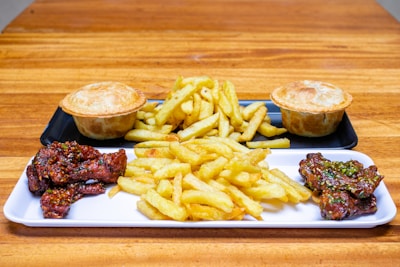 A wooden table holds a serving platter featuring crispy fries in the center, flanked by two small meat pies. Adjacent to the fries are portions of glazed chicken wings, sprinkled with herbs and sesame seeds. The food items rest on rectangular serving trays.