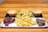 A wooden table holds a serving platter featuring crispy fries in the center, flanked by two small meat pies. Adjacent to the fries are portions of glazed chicken wings, sprinkled with herbs and sesame seeds. The food items rest on rectangular serving trays.