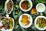An array of dishes featuring various foods is presented on a dark surface scattered with rice grains. The dishes include grilled fish garnished with slices of tomato and onion, a richly spiced meat stew with vegetables, a plate featuring fried eggs and cut vegetables, and a dish containing chapati with leafy greens and sliced tomatoes. Fresh ingredients such as a whole lime, an onion, ginger, a coconut, and various chilies accompany the setup, adding vibrant colors and fresh appeal.