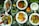 An array of dishes featuring various foods is presented on a dark surface scattered with rice grains. The dishes include grilled fish garnished with slices of tomato and onion, a richly spiced meat stew with vegetables, a plate featuring fried eggs and cut vegetables, and a dish containing chapati with leafy greens and sliced tomatoes. Fresh ingredients such as a whole lime, an onion, ginger, a coconut, and various chilies accompany the setup, adding vibrant colors and fresh appeal.