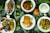 An array of dishes featuring various foods is presented on a dark surface scattered with rice grains. The dishes include grilled fish garnished with slices of tomato and onion, a richly spiced meat stew with vegetables, a plate featuring fried eggs and cut vegetables, and a dish containing chapati with leafy greens and sliced tomatoes. Fresh ingredients such as a whole lime, an onion, ginger, a coconut, and various chilies accompany the setup, adding vibrant colors and fresh appeal.