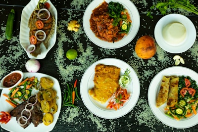 An array of dishes featuring various foods is presented on a dark surface scattered with rice grains. The dishes include grilled fish garnished with slices of tomato and onion, a richly spiced meat stew with vegetables, a plate featuring fried eggs and cut vegetables, and a dish containing chapati with leafy greens and sliced tomatoes. Fresh ingredients such as a whole lime, an onion, ginger, a coconut, and various chilies accompany the setup, adding vibrant colors and fresh appeal.