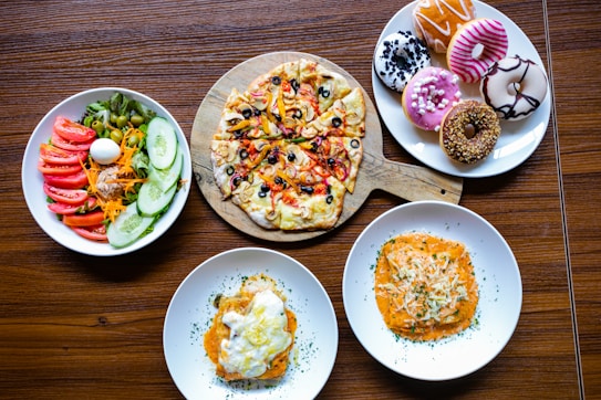 A variety of dishes including a colorful vegetable salad with sliced cucumber, tomato, olives, grated carrot, and a boiled egg are arranged on a plate. Next to it is a wooden board holding a pizza topped with cheese, olives, bell peppers, and onions. Another plate holds an assortment of decorated donuts in different colors and toppings. Two separate plates each contain a serving of pasta with creamy sauces, garnished with herbs and cheese.
