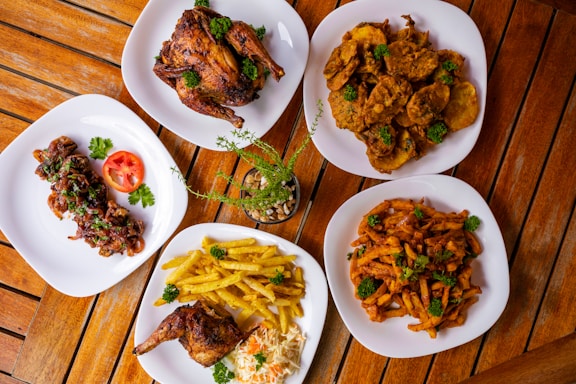 Several plates of food are arranged on a wooden table, featuring roasted chicken, fried potatoes, sautéed plantains, grilled chicken skewers, and sauced fries. Garnishes of parsley add a touch of green, with a small potted plant in the center.