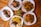 Several plates of food are arranged on a wooden table, featuring roasted chicken, fried potatoes, sautéed plantains, grilled chicken skewers, and sauced fries. Garnishes of parsley add a touch of green, with a small potted plant in the center.