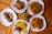 Several plates of food are arranged on a wooden table, featuring roasted chicken, fried potatoes, sautéed plantains, grilled chicken skewers, and sauced fries. Garnishes of parsley add a touch of green, with a small potted plant in the center.