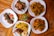 Several plates of food are arranged on a wooden table, featuring roasted chicken, fried potatoes, sautéed plantains, grilled chicken skewers, and sauced fries. Garnishes of parsley add a touch of green, with a small potted plant in the center.