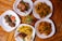 Several plates of food are arranged on a wooden table, featuring roasted chicken, fried potatoes, sautéed plantains, grilled chicken skewers, and sauced fries. Garnishes of parsley add a touch of green, with a small potted plant in the center.