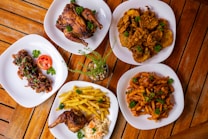 Several plates of food are arranged on a wooden table, featuring roasted chicken, fried potatoes, saut&eacute;ed plantains, grilled chicken skewers, and sauced fries. Garnishes of parsley add a touch of green, with a small potted plant in the center.