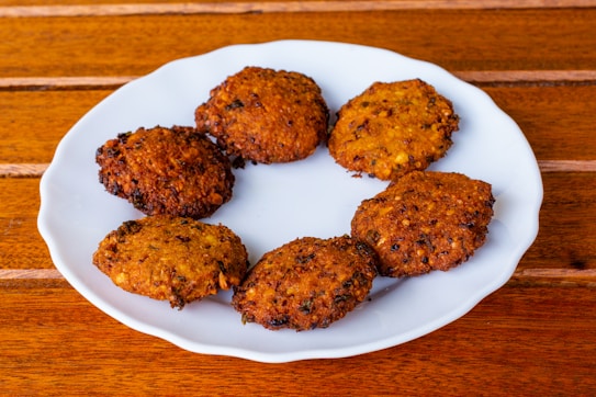 A white plate contains seven round, crispy, brown fritters arranged in a circular pattern. The fritters have a textured surface, indicating a deep-fried preparation, and a few visible herbs or spices mixed within. This setup is placed on a wooden surface, adding a rustic quality to the scene.