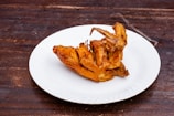 A piece of fried chicken is placed on a white plate. The background is a dark wooden surface, providing a rustic contrast to the golden-brown color of the chicken. The chicken appears crispy and well-cooked, with visible texture on the skin.
