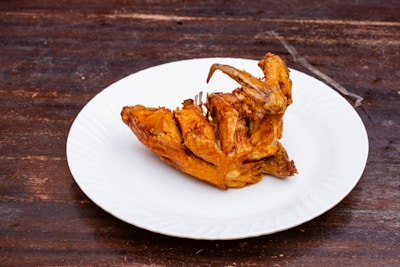 A piece of fried chicken is placed on a white plate. The background is a dark wooden surface, providing a rustic contrast to the golden-brown color of the chicken. The chicken appears crispy and well-cooked, with visible texture on the skin.