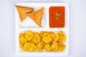 A white divided plate contains two samosas, a dish of red dipping sauce garnished with green herbs, and a serving of fried potato slices.