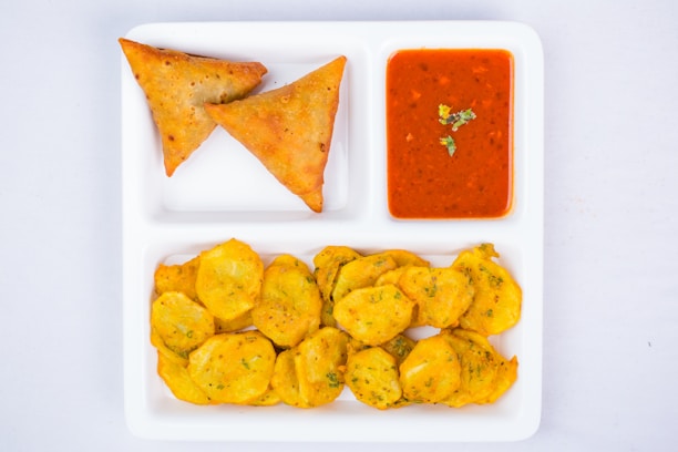 A white divided plate contains two samosas, a dish of red dipping sauce garnished with green herbs, and a serving of fried potato slices.