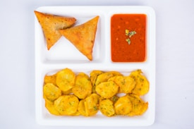 A white divided plate contains two samosas, a dish of red dipping sauce garnished with green herbs, and a serving of fried potato slices.