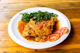 A white plate containing a serving of chicken stew garnished with vibrant red tomatoes and accompanied by cooked green leafy vegetables. The dish is placed on a wooden surface, adding a rustic touch to the presentation.