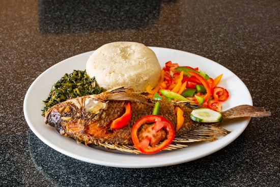 A deliciously prepared grilled fish served on a white plate, accompanied by leafy greens, a portion of boiled maize dough, and a colorful array of sliced vegetables, including bell peppers and tomatoes.